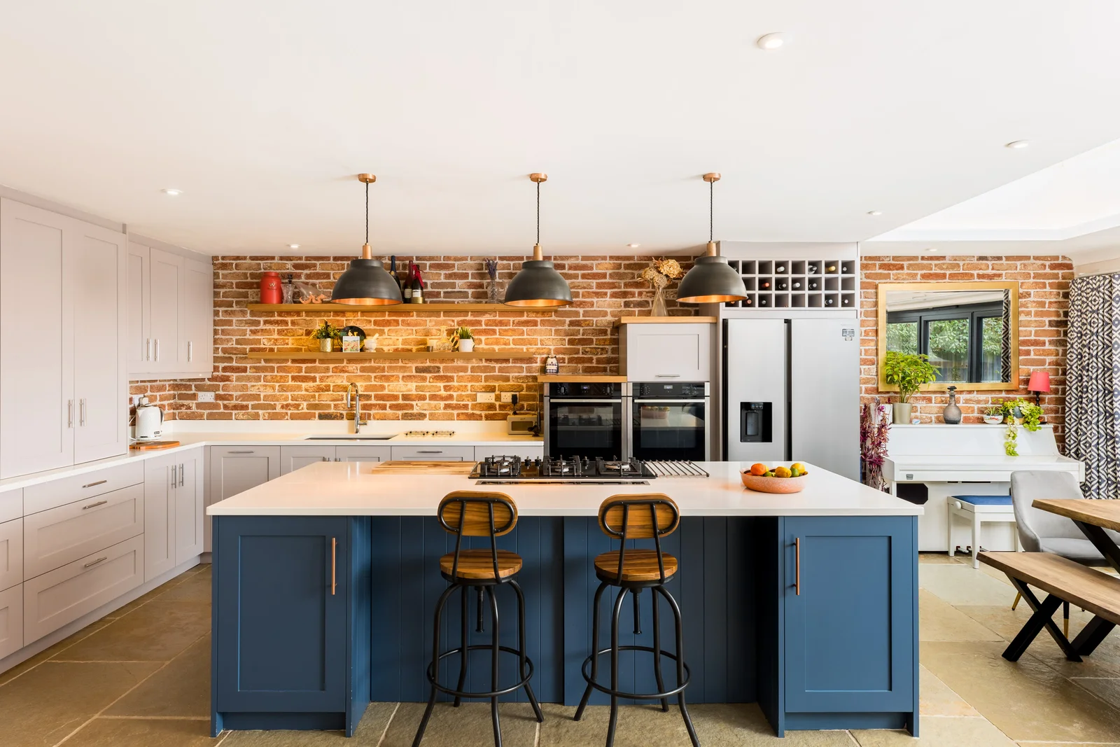 Kitchen with exposed brick wall and navy island under brass pendants